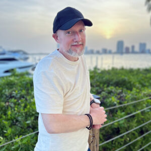 A man with a light beard, wearing a black cap and a beige T-shirt, stands near a waterfront railing at sunset. He is smiling gently, with a city skyline and a yacht in the background.